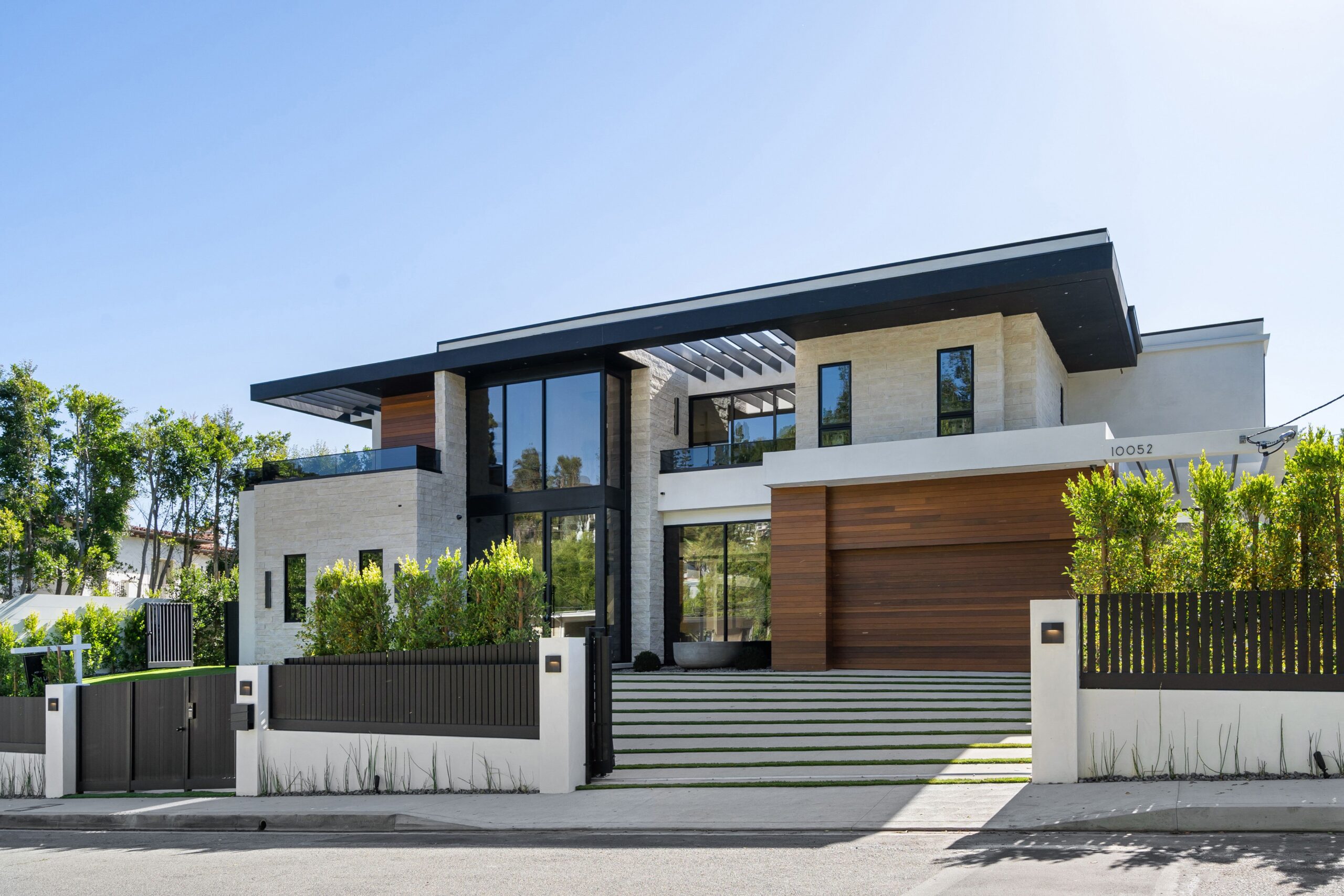 Modern luxury house with large windows and a wooden garage door, surrounded by greenery and a clear blue sky.